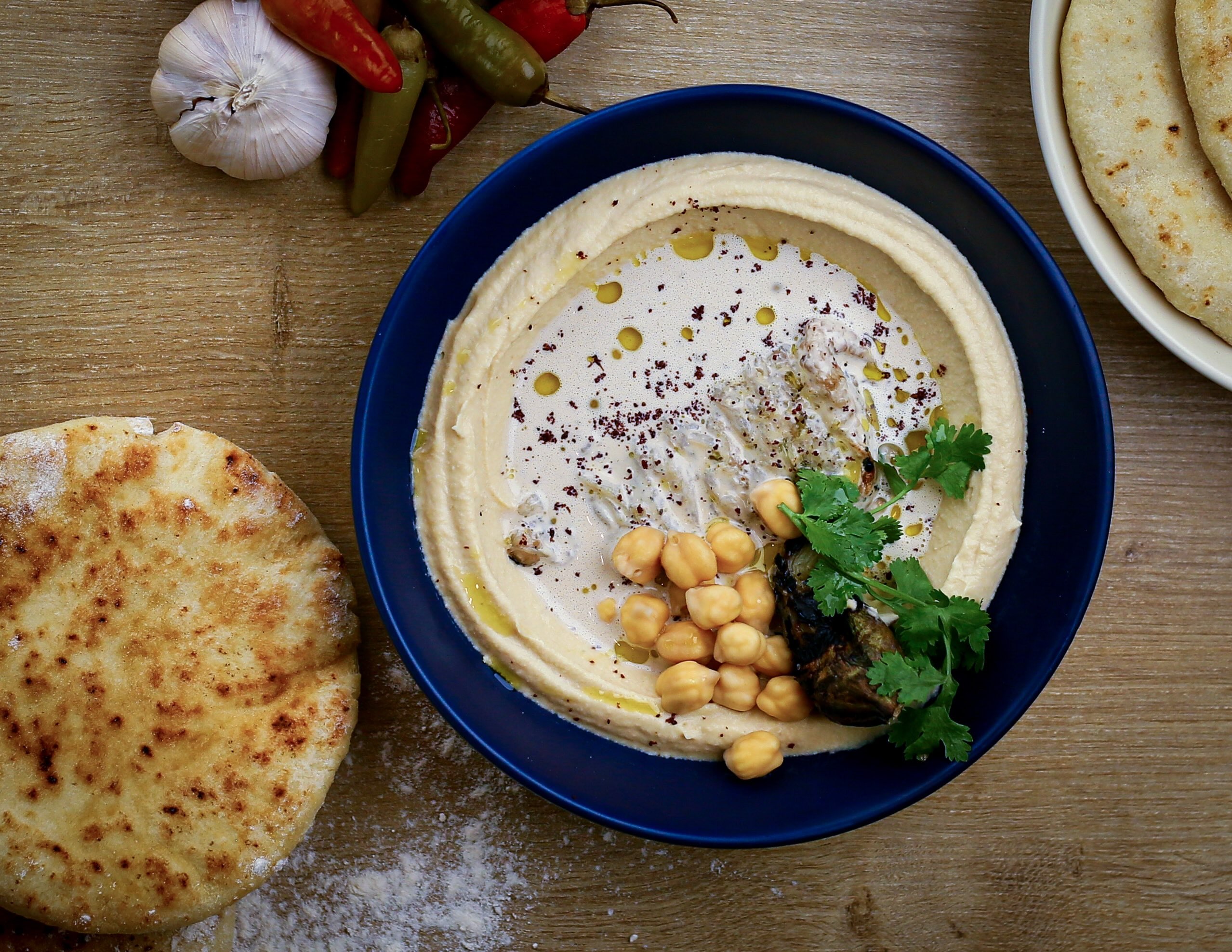 Birds-eye view photo of a wood table with a blue bowl of hummus and some chickpeas. One flat bread, garlic and peppers.