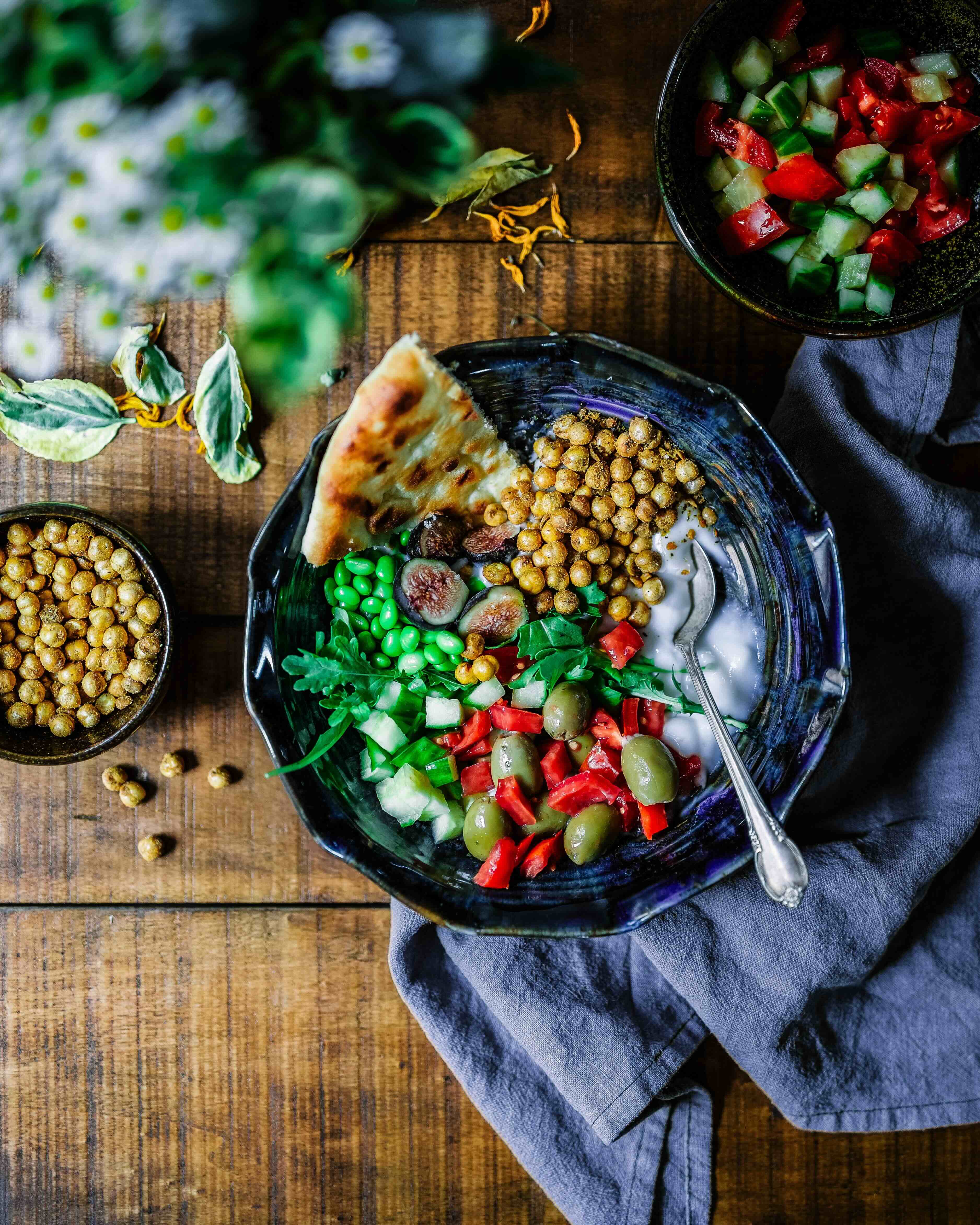 Birds-eye view photo of a wood table with a blue-grey kitchen towel under one blue bowl with chickpeas, yoghurt, peppers and olives, edamame, fig and a piece of a flat bread. On the right side of it, there is a bowl with chopped cucumber and tomatoes. And on the left side there is a bowl with more chickpeas.