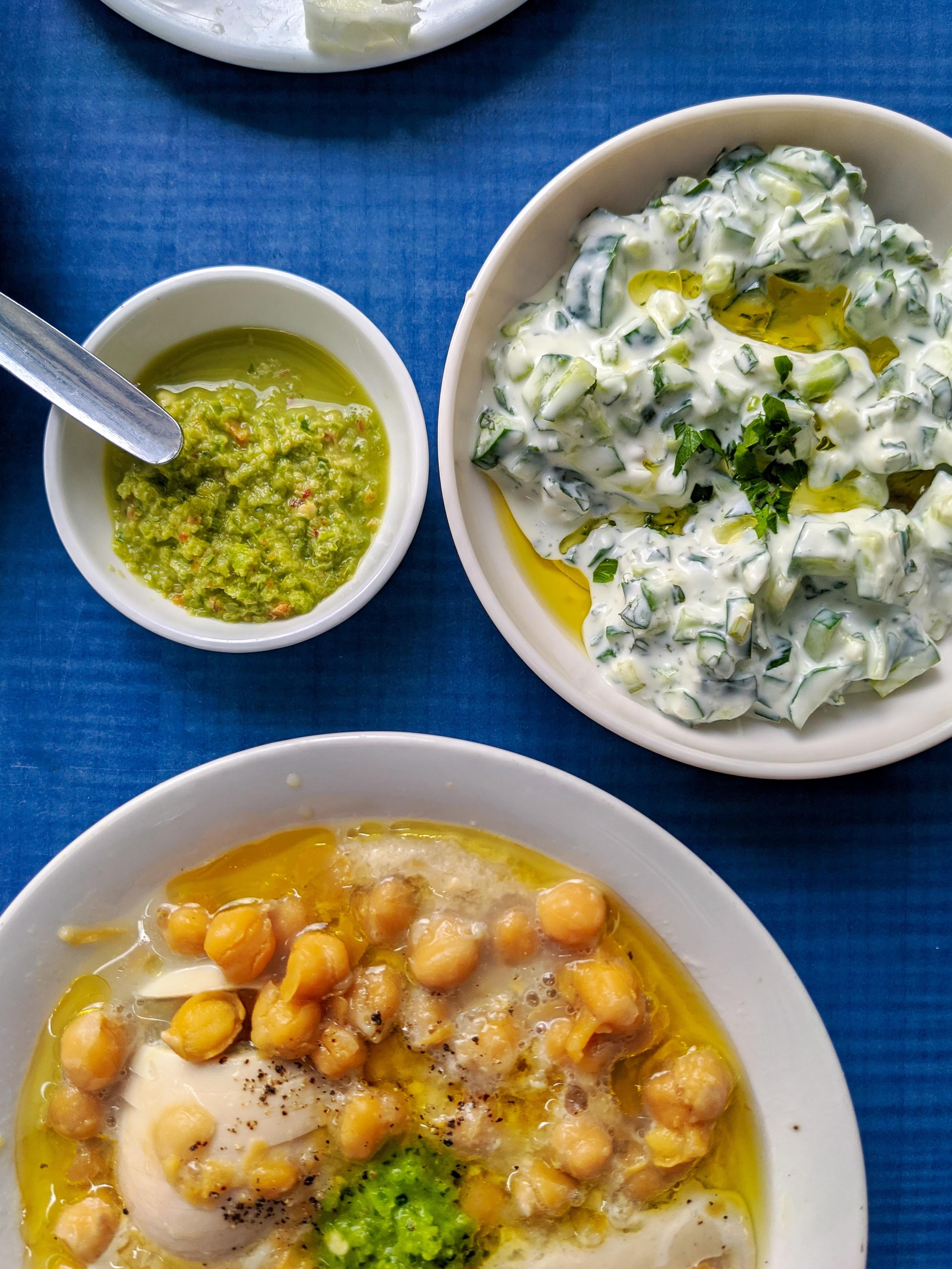 Birds-eye view photo of a blue table with one plate of chickpeas, one bowl with tzatsiki and one smaller bowl with som green mushy stuff.